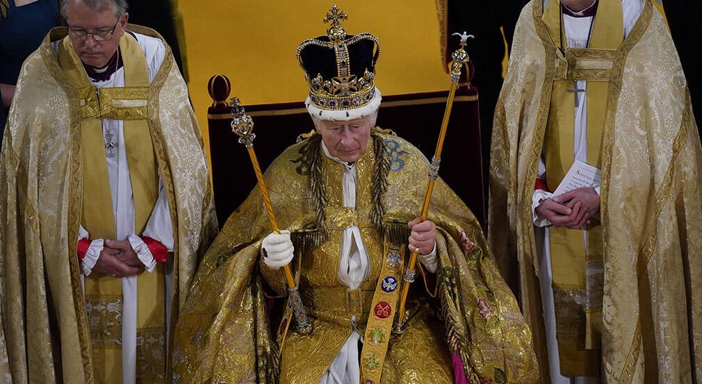 King Charles III seated wearing a golden robes and wearing a gold crown. He is holding 2 gold rods. 