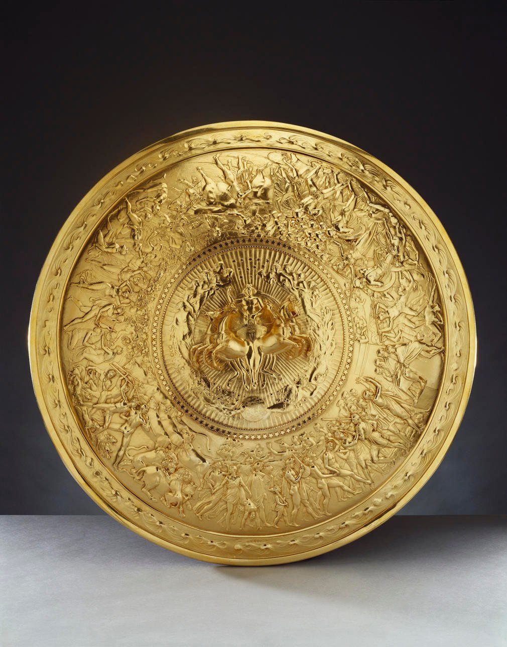 A silver-gilt convex shield with a central medallion cast in high relief showing Apollo in a quadriga, surrounded by stars and female figures representing the constellations. The broad border is cast in low relief with scenes of human life (a wedding and 