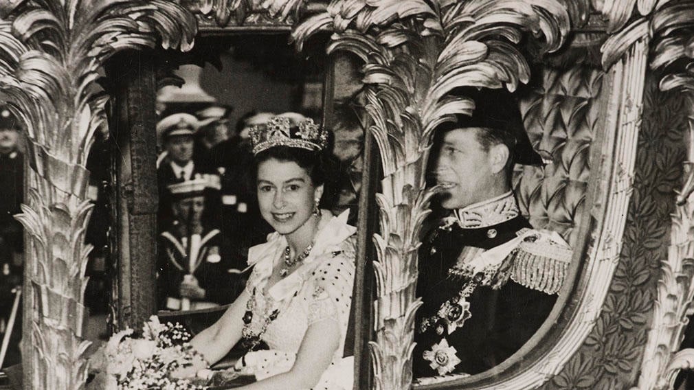 Photograph of HM Queen Elizabeth II sitting beside HRH The Prince Philip, Duke of Edinburgh in the Gold State Coach on their way to Westminster Abbey for the Coronation Service. In the background can be seen Naval Officers with rifles and Police