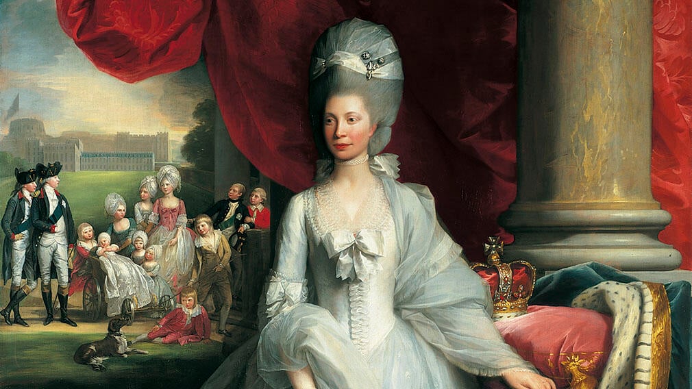 Queen Charlotte standing wearing a white dress with bows on the front. Her grey hair in a tall oval shaped hairstyle. A crown sits on a table beside her.