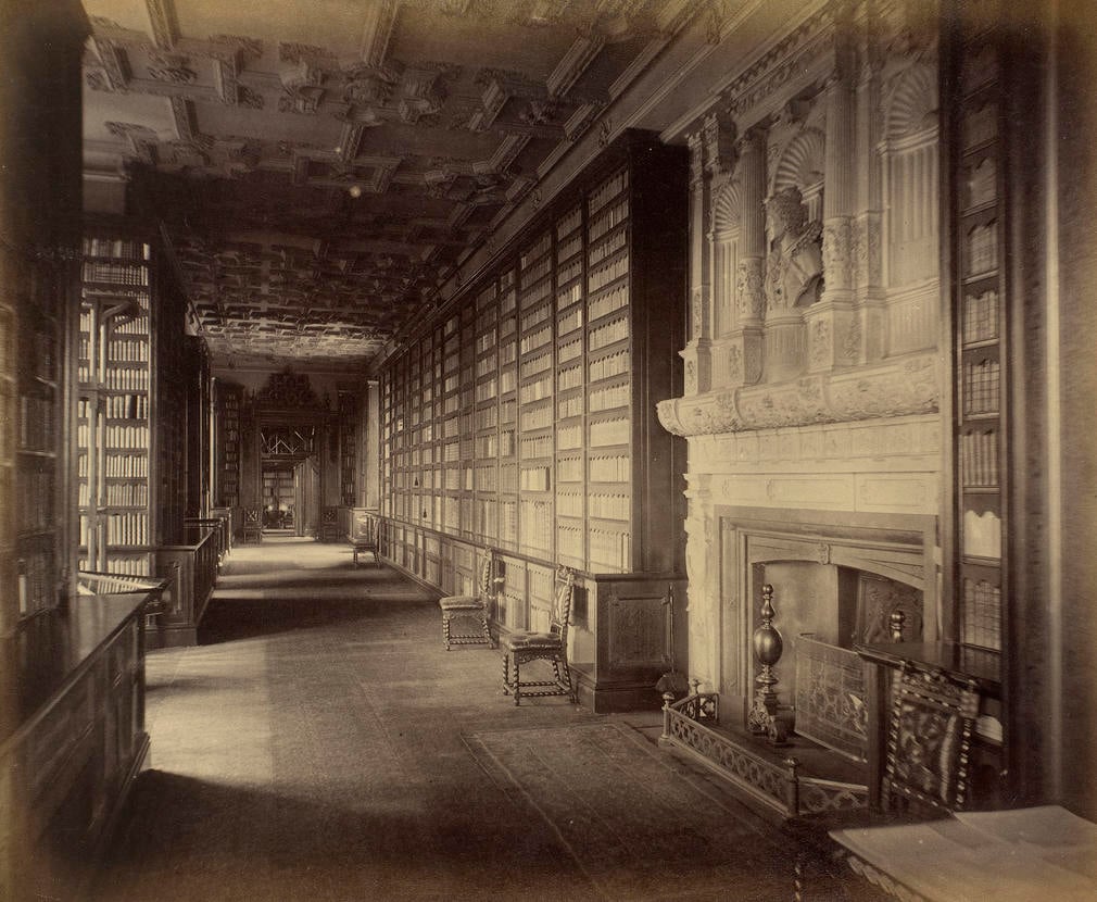 Queen Elizabeth's Gallery, in the Royal Library, WC. Shelves filled with books are either side of the walkway, chairs stand to the right. Window bays are on the left & a fireplace with carved niches and a bust above are (near) right.