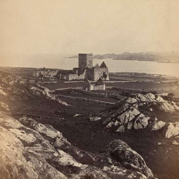 Photograph of the ruins of Iona Abbey situated on the Ross of Mull, a large peninsula on the Isle of Mull. In the foreground are large boulders. The central tower of the Abbey stands above the ruins of the nave and another ruined, smaller dwelling st