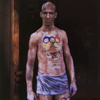 Colour photograph of a three-quarter length portrait of a man standing, naked apart from a blue coloured piece of cloth tied around his hips. He faces the camera front on, staring down the camera lens. He is covered is white and black paint and 