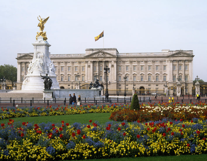 The grey stone facade of Buckingham Palace with gates and flower beds