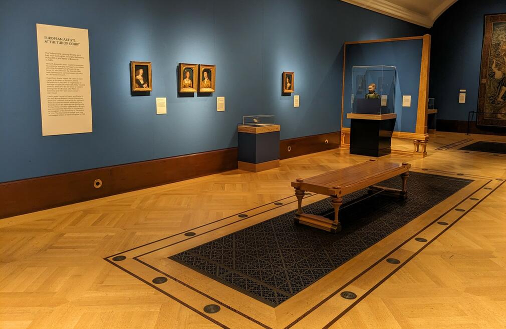 A gallery space with blue walls, wooden floor and metal grill. There are paintings on the walls.