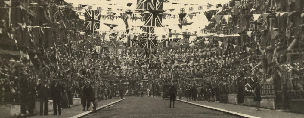 Black and white photo showing a street decorated with large amounts of flags and bunting