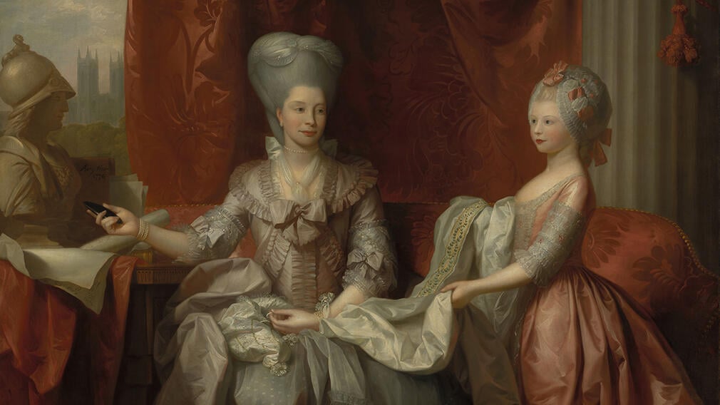 Two women wearing elaborate gowns are embroidering a piece of material. Queen Charlotte on the left has grey hair in a tall style and is wearing a light pink dress.