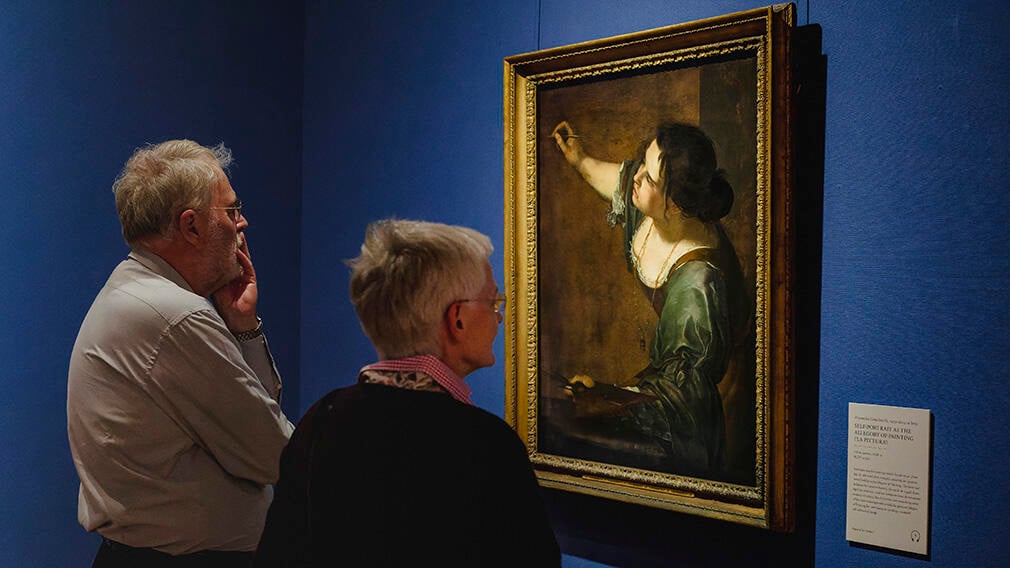 A man and a woman looking at a painting hanging on the wall. They both have grey hair.