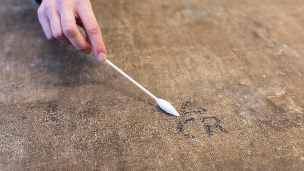 A person holding a cotton bud cleans the surface of the back of a canvas. 