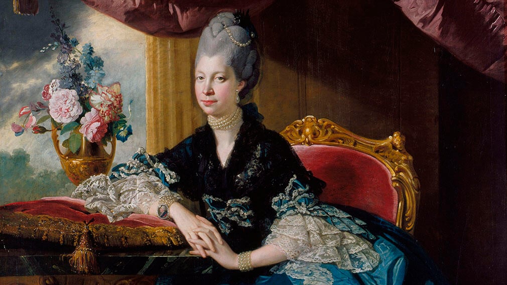 Queen Charlotte sitting on a red velvet chair wearing a blue silk dress with white lace. She also wears a black shawl. A vase of flowers sits on a table on her left.