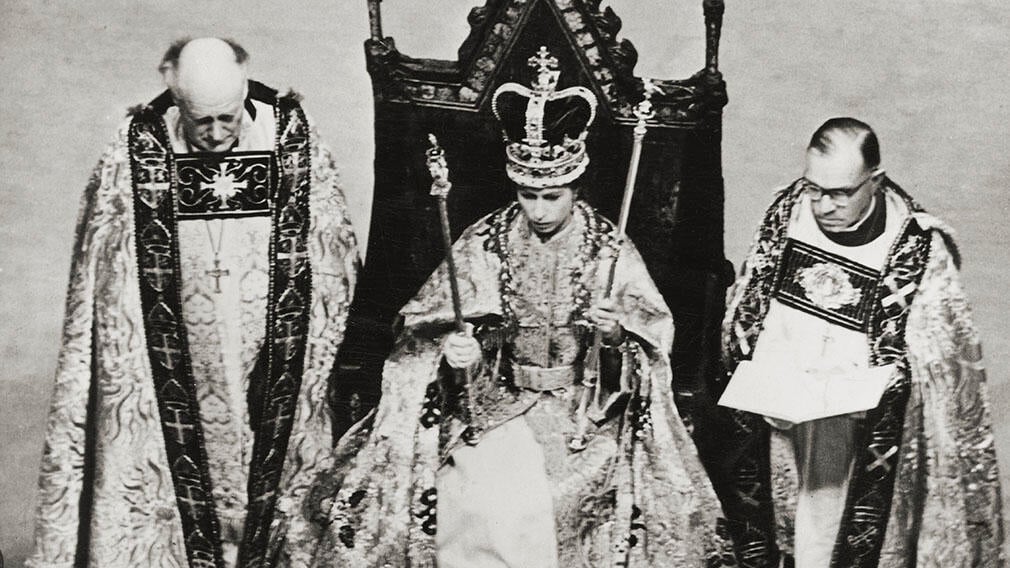 Black and white photograph of Queen Elizabeth II seated on the Coronation Chair in Westminster Abbey. She is holding two sceptres and wearing a crown. 