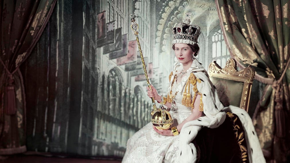 Photograph of Queen Elizabeth II seated wearing coronation robes. She poses against a backdrop depicting the interior of Westminster Abbey