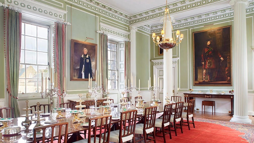 A lavish dining room with the table laid and decorated. The room is filled with paintings and a chandelier.