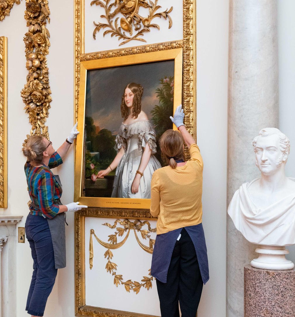 Two technicians hang one of the cleaned and conserved paintings in the Marble Hall, Buckingham Palace